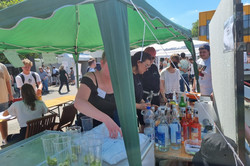 Ein grüner, leicht schiefer Pavillon spendet in diesem Bild einigen Menschen Schatten. Zwei Tische, eine große Kühltruhe, ein Einkaufswagen voller Getränke und einige Becher mit Limetten finden ebenfalls Schatten unter dem Pavillon. Das kann nur unser geliebter Cocktailstand sein! Und er ist es auch. Bei hellblauem Himmel und einer Menge Menschen auf dem Campus lässt sich ein Cocktail schließlich auch super genießen!