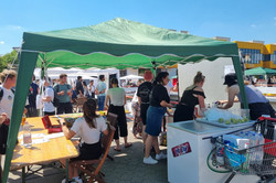 Ein grüner, leicht schiefer Pavillon spendet in diesem Bild einigen Menschen Schatten. Zwei Tische, eine große Kühltruhe, ein Einkaufswagen voller Getränke und einige Becher mit Limetten finden ebenfalls Schatten unter dem Pavillon. Das kann nur unser geliebter Cocktailstand sein! Und er ist es auch. Bei hellblauem Himmel und einer Menge Menschen auf dem Campus lässt sich ein Cocktail schließlich auch super genießen!