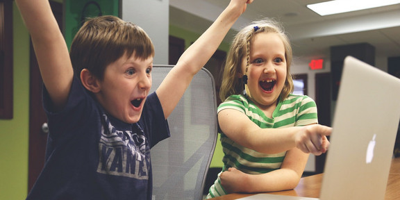 Zwei Kinder, ein Junge und ein Mädchen (ca. Grundschulalter) sitzen vor einem Macbook und freuen sich. Der Junge hebt jubelnd seine Arme, während das Mädchen auf das geöffnete MacBook zeigt.