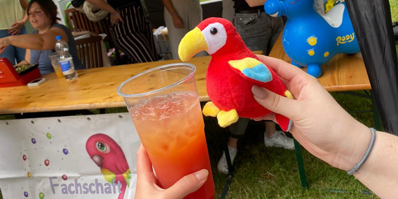 Das Stofftier der Fachschaft - ein roter Papagei - trinkt aus einem orangenen Cocktail. Im Hintergrund ist der Cocktailstand der Fachschaft zu erkennen.