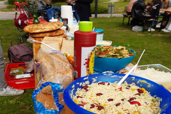 Wer sich dieses Bild ansieht bekommt sofort Hunger. Im Zentrum stehen nämlich allerlei Salate, Saucen, Brote und noch viele weitere Leckereien. Etwas weiter im Hintergrund sind ein paar Studierende zu erahnen, die sich hungrig auf die Leckereien stürzen. 