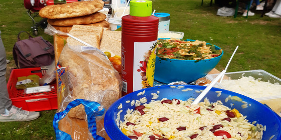 Wer sich dieses Bild ansieht bekommt sofort Hunger. Im Zentrum stehen nämlich allerlei Salate, Saucen, Brote und noch viele weitere Leckereien. Etwas weiter im Hintergrund sind ein paar Studierende zu erahnen, die sich hungrig auf die Leckereien stürzen. 