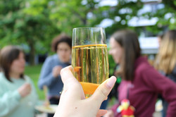 Ein Glas Sekt wird in die Kamera gehalten. Im Hintergrund unterhalten sich drei Frauen.
