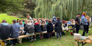 Auf dem Bild sind viele Menschen erkennbar. Sie sitzen auf Bierbänken auf einer Wiese. Es ist eine Grillveranstaltung aus 2022.