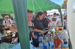 Ein grüner, leicht schiefer Pavillon spendet in diesem Bild einigen Menschen Schatten. Zwei Tische, eine große Kühltruhe, ein Einkaufswagen voller Getränke und einige Becher mit Limetten finden ebenfalls Schatten unter dem Pavillon. Das kann nur unser geliebter Cocktailstand sein! Und er ist es auch. Bei hellblauem Himmel und einer Menge Menschen auf dem Campus lässt sich ein Cocktail schließlich auch super genießen!