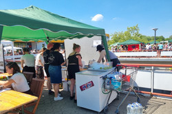 Ein grüner, leicht schiefer Pavillon spendet in diesem Bild einigen Menschen Schatten. Zwei Tische, eine große Kühltruhe, ein Einkaufswagen voller Getränke und einige Becher mit Limetten finden ebenfalls Schatten unter dem Pavillon. Das kann nur unser geliebter Cocktailstand sein! Und er ist es auch. Bei hellblauem Himmel und einer Menge Menschen auf dem Campus lässt sich ein Cocktail schließlich auch super genießen!